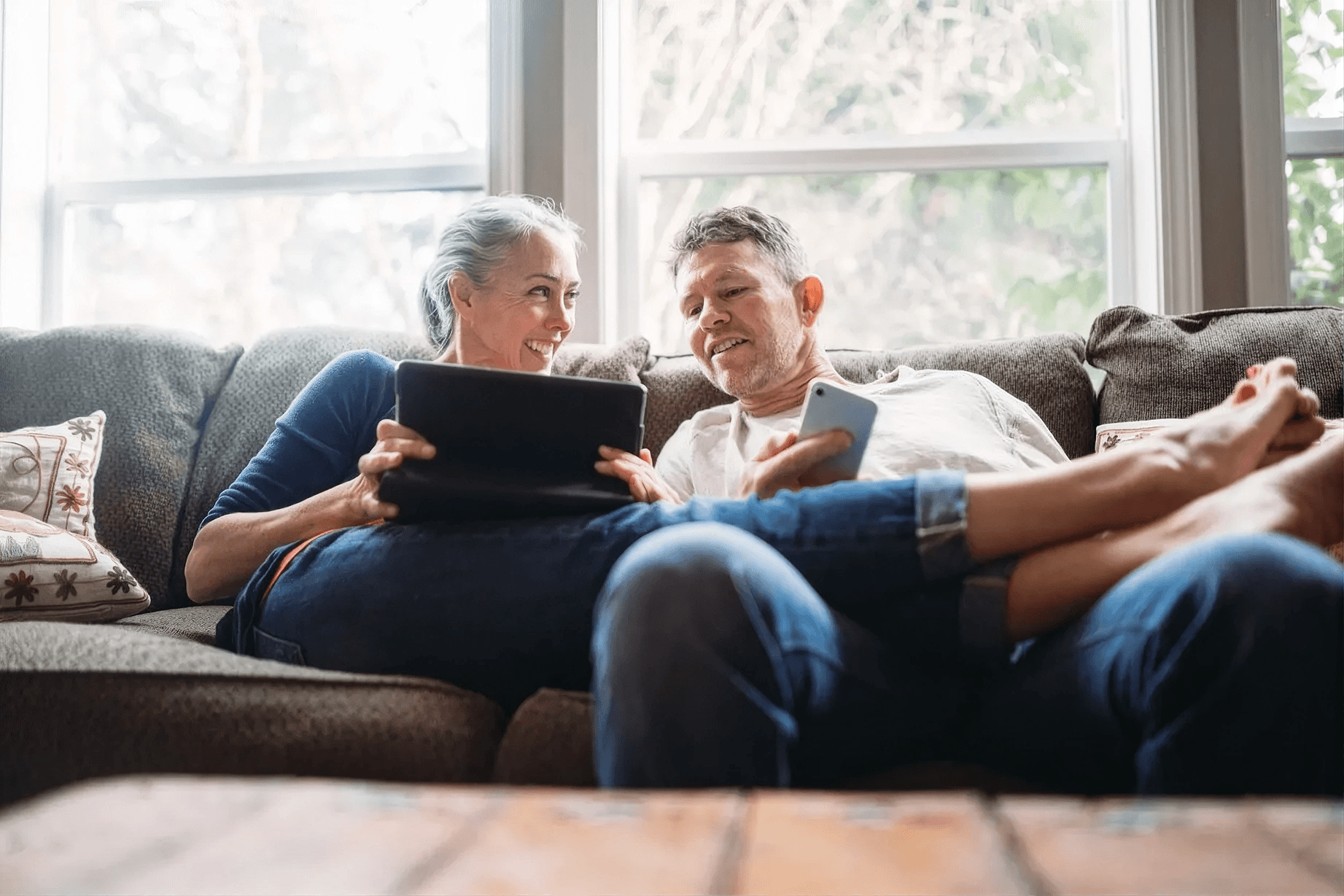 Smiling couple relaxing together on a living room sofa while using a tablet and smartphone, enjoying the comfort and peace of mind that comes from a safe, well-maintained home with reliable plumbing and water systems.