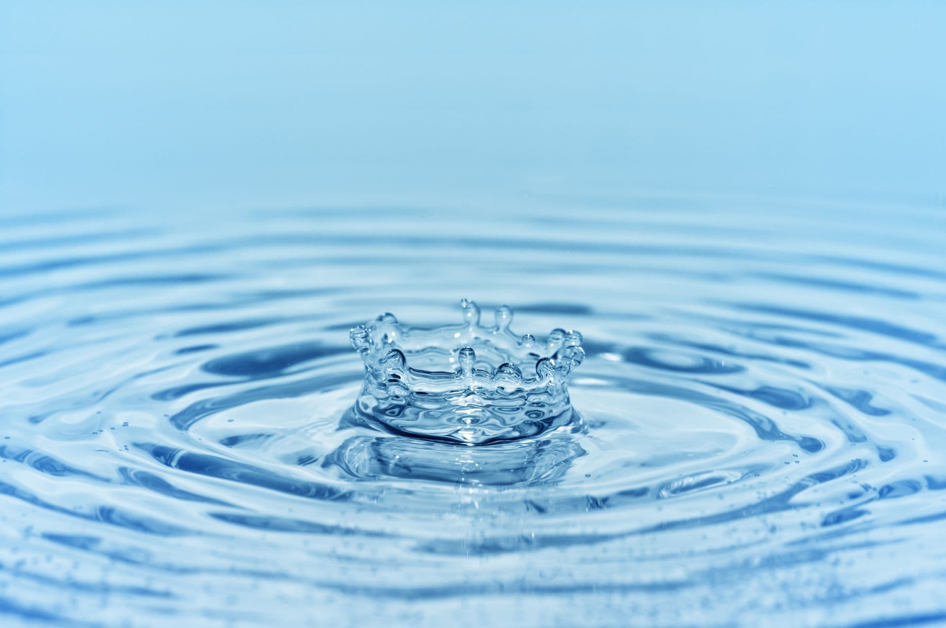 Close-up of a single water droplet creating a crown-shaped splash on a calm blue water surface, with concentric ripples radiating outward against a soft gradient background, symbolizing clean, pure, and refreshing water.