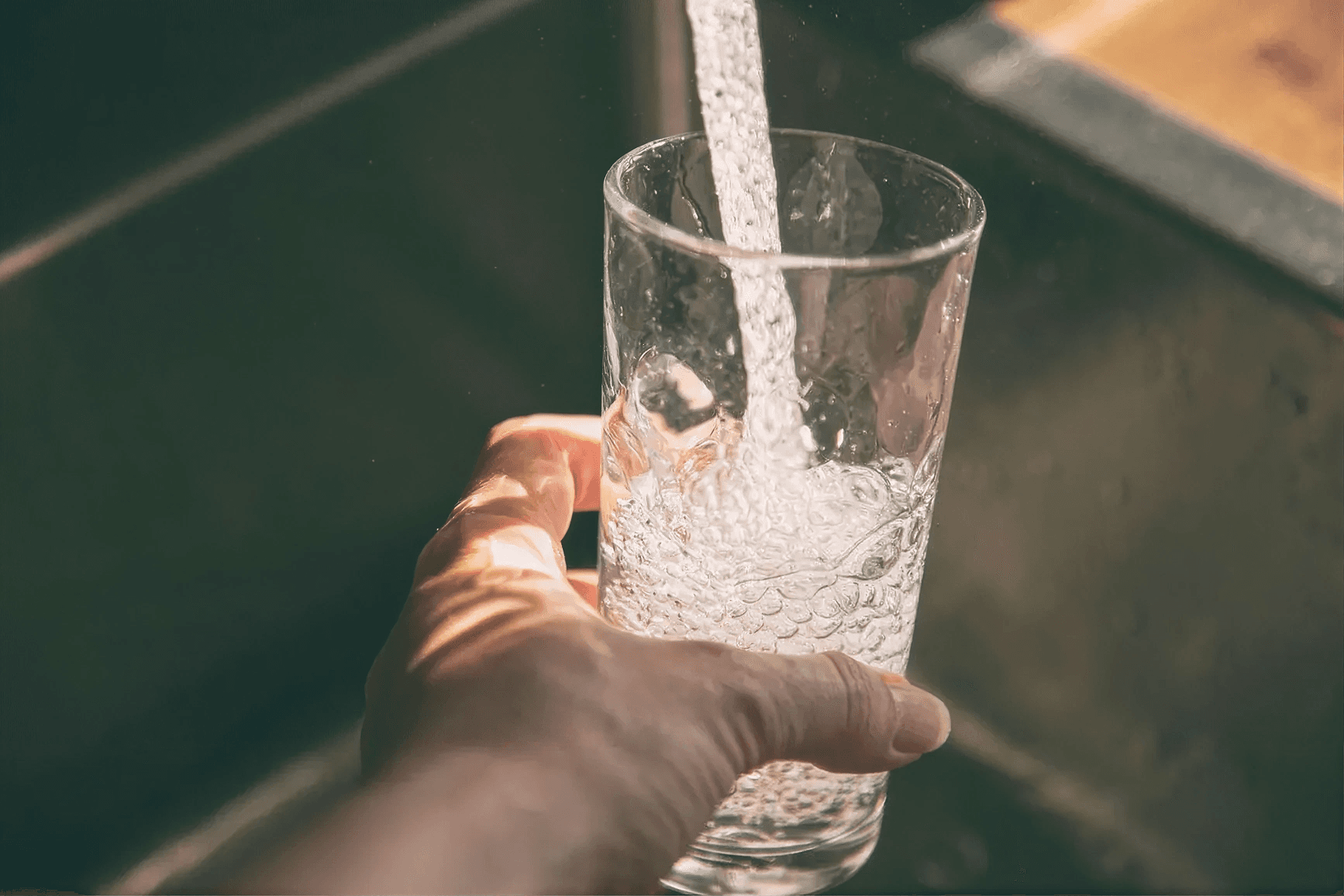 Hand holding a clear drinking glass while filling it with fresh, clean water from a kitchen faucet, showcasing filtered drinking water in a residential home.