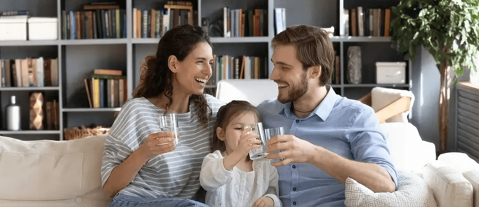 Smiling family sitting together on a living room sofa, each holding a glass of clean drinking water, highlighting healthy hydration and access to filtered water at home.