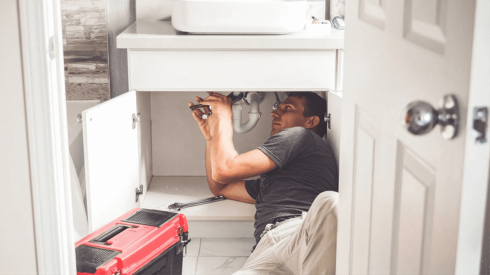 Professional plumber repairing plumbing beneath a bathroom vanity sink, using hand tools to inspect and service drain pipes and water supply connections during a residential plumbing repair.