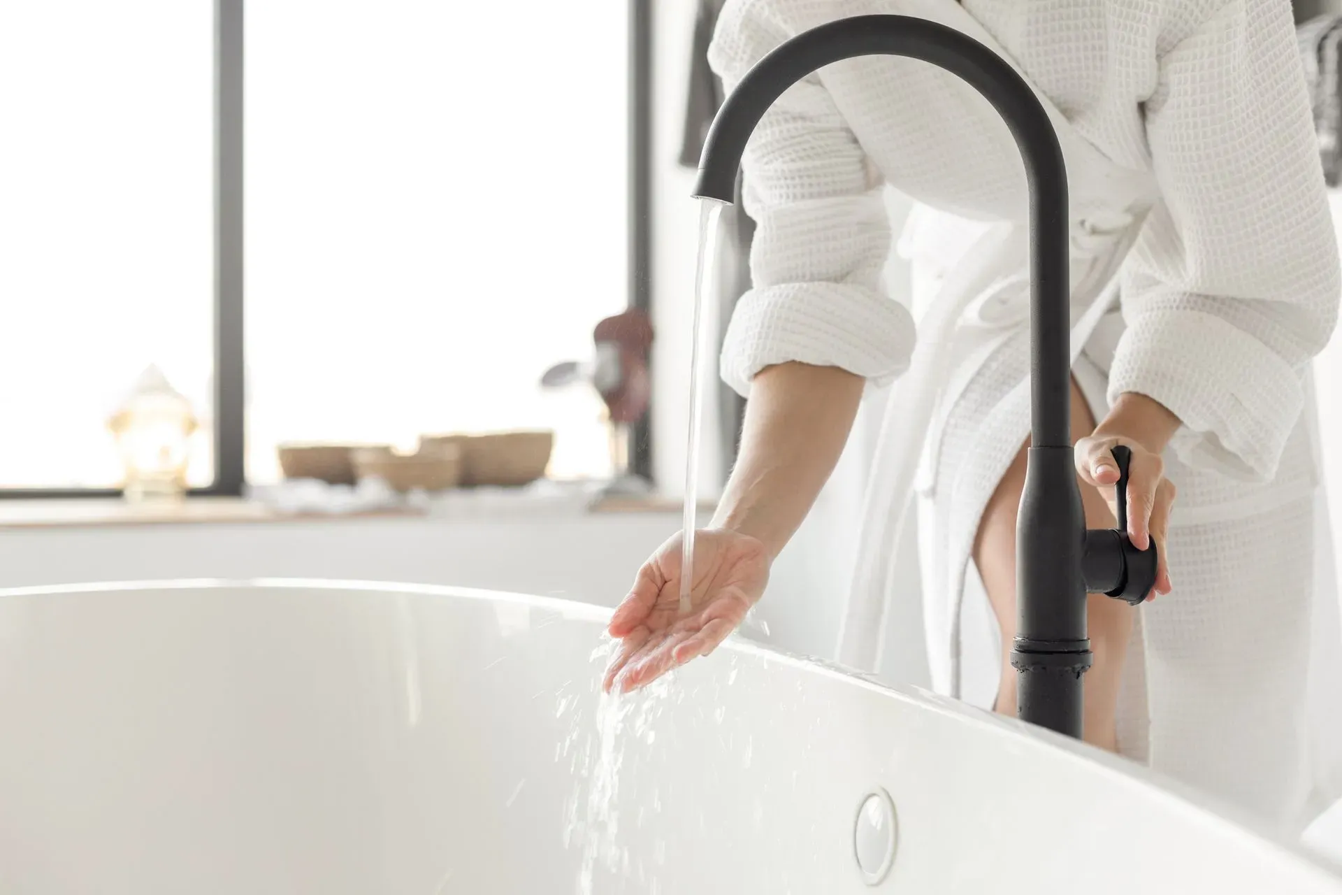 Modern freestanding bathtub being filled with water from a matte black tub faucet while a person in a white bathrobe checks the water temperature before bathing.